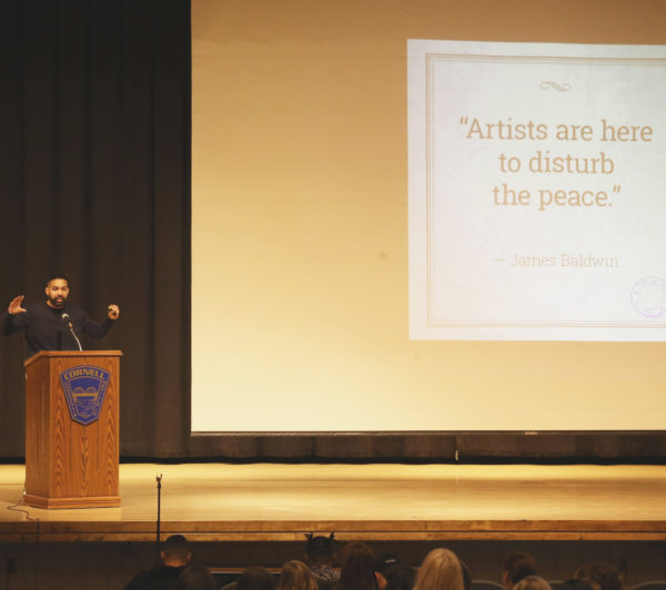 Jasiri X gives a speech in front of a screen reading "Artists are here to disturb the peace."