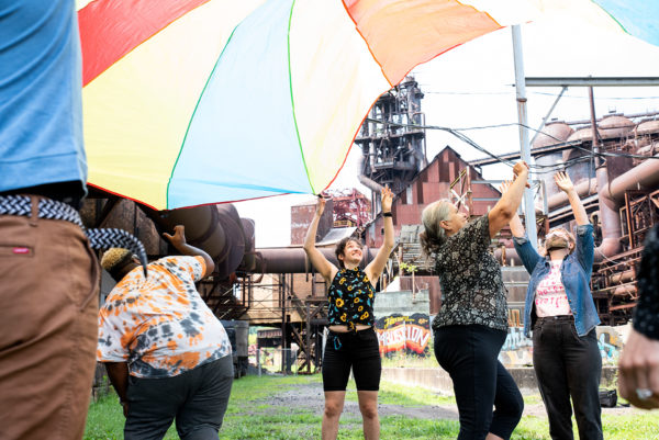 Arts-Ed-Collab_July-2020_WEB_065 A diverse group of educators laughs beneath a large colorful parachute at an industrial site.