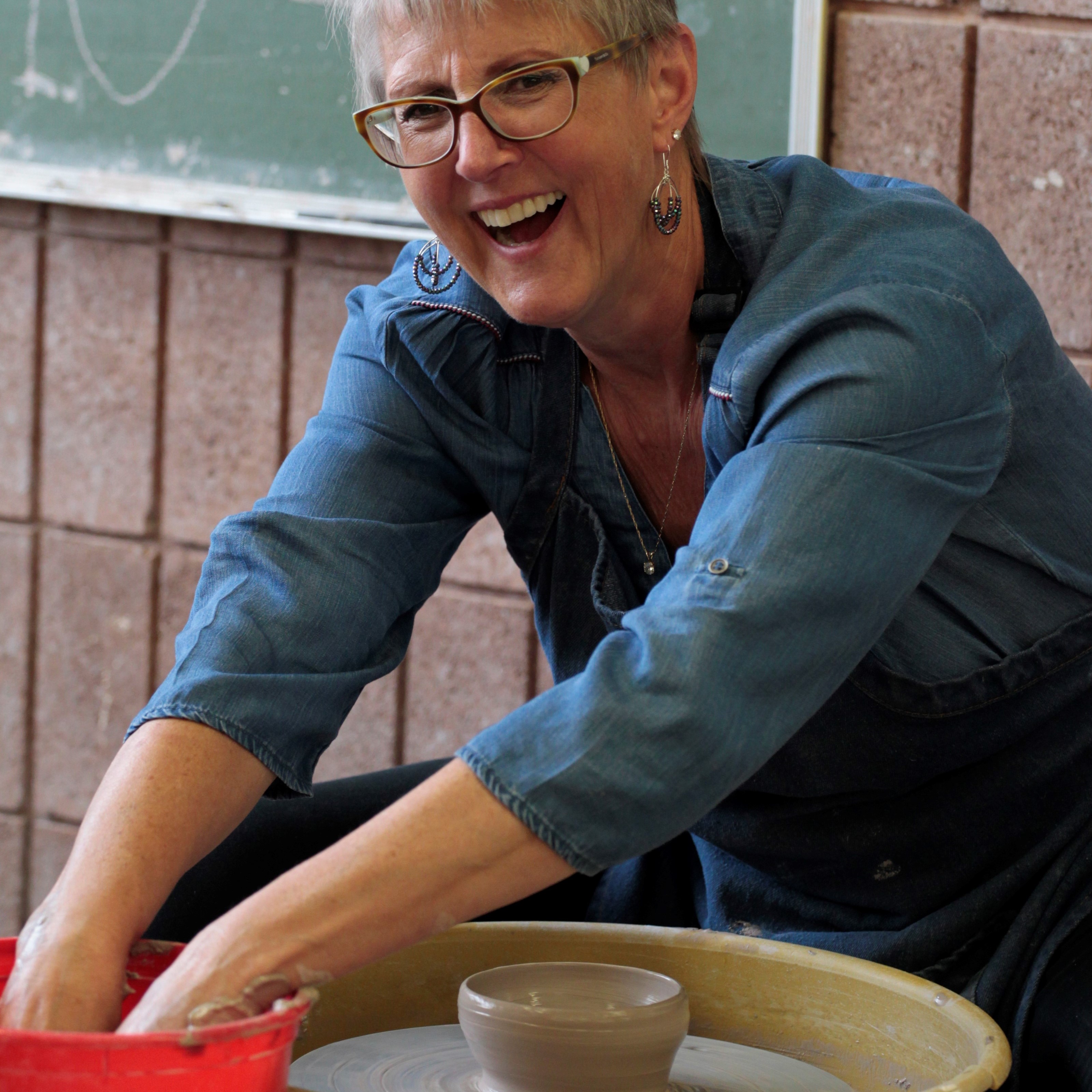 A woman laughs while working with wet clay on a potter's wheel.