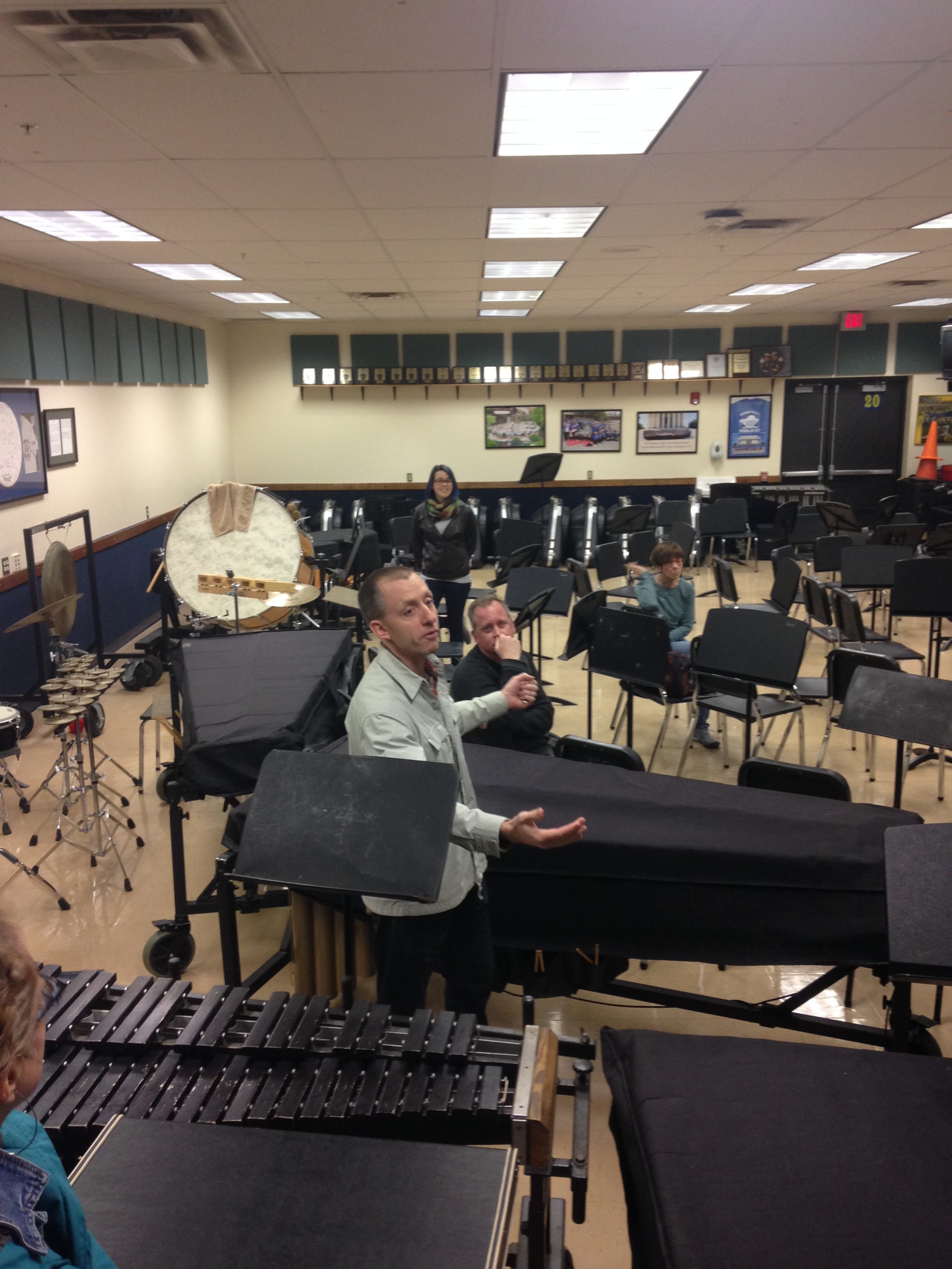 A band instructor discusses Hampton Township's band program in a large band room full of music stands and instruments.