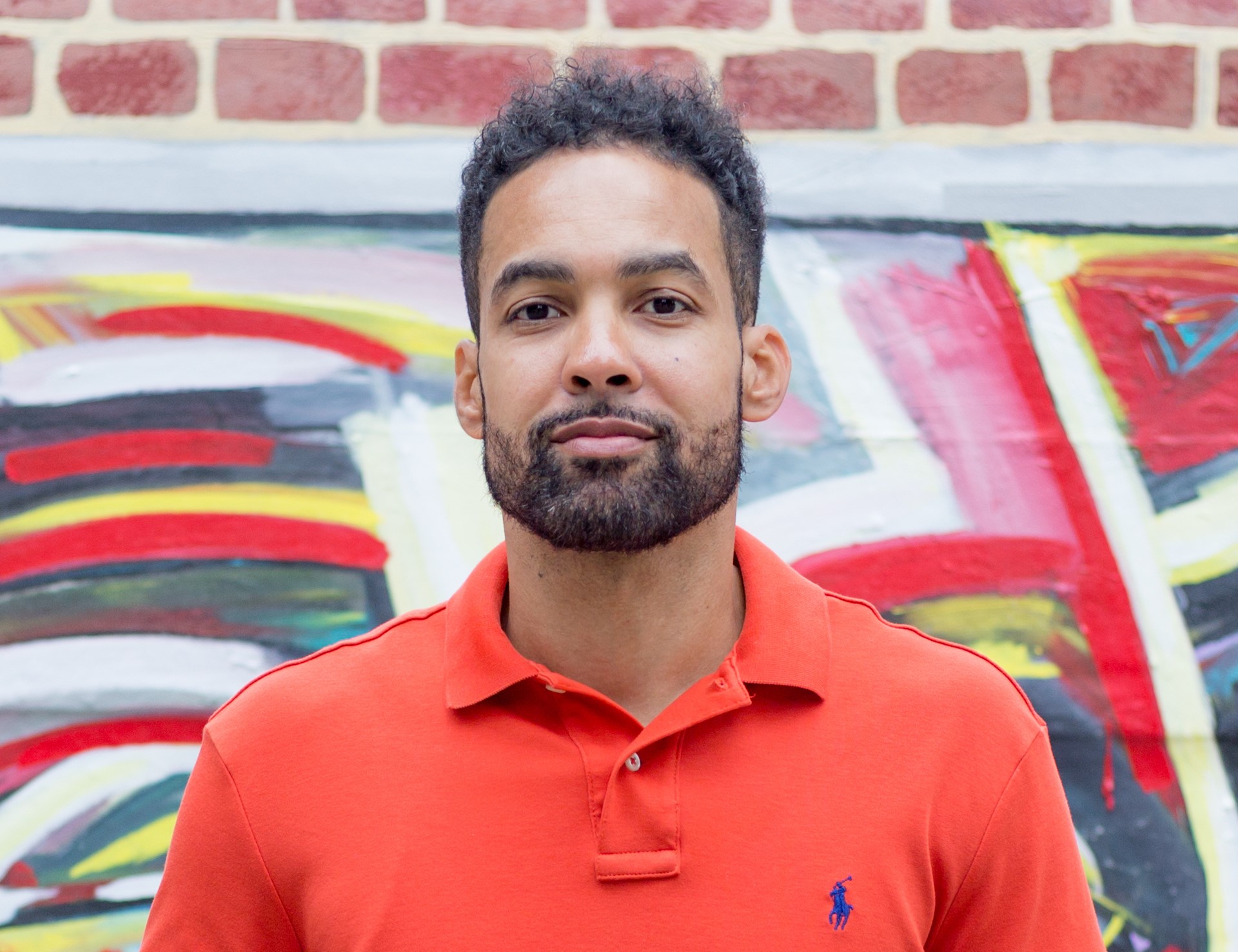 Jasiri X in a polo shirt smiling in front of a brightly painted brick wall.