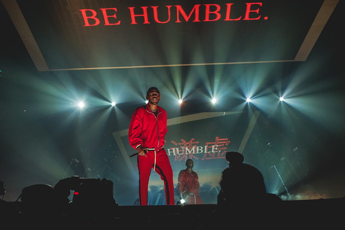 Kendrick Lamar holding a microphone onstage beneath the words "Be Humble."
