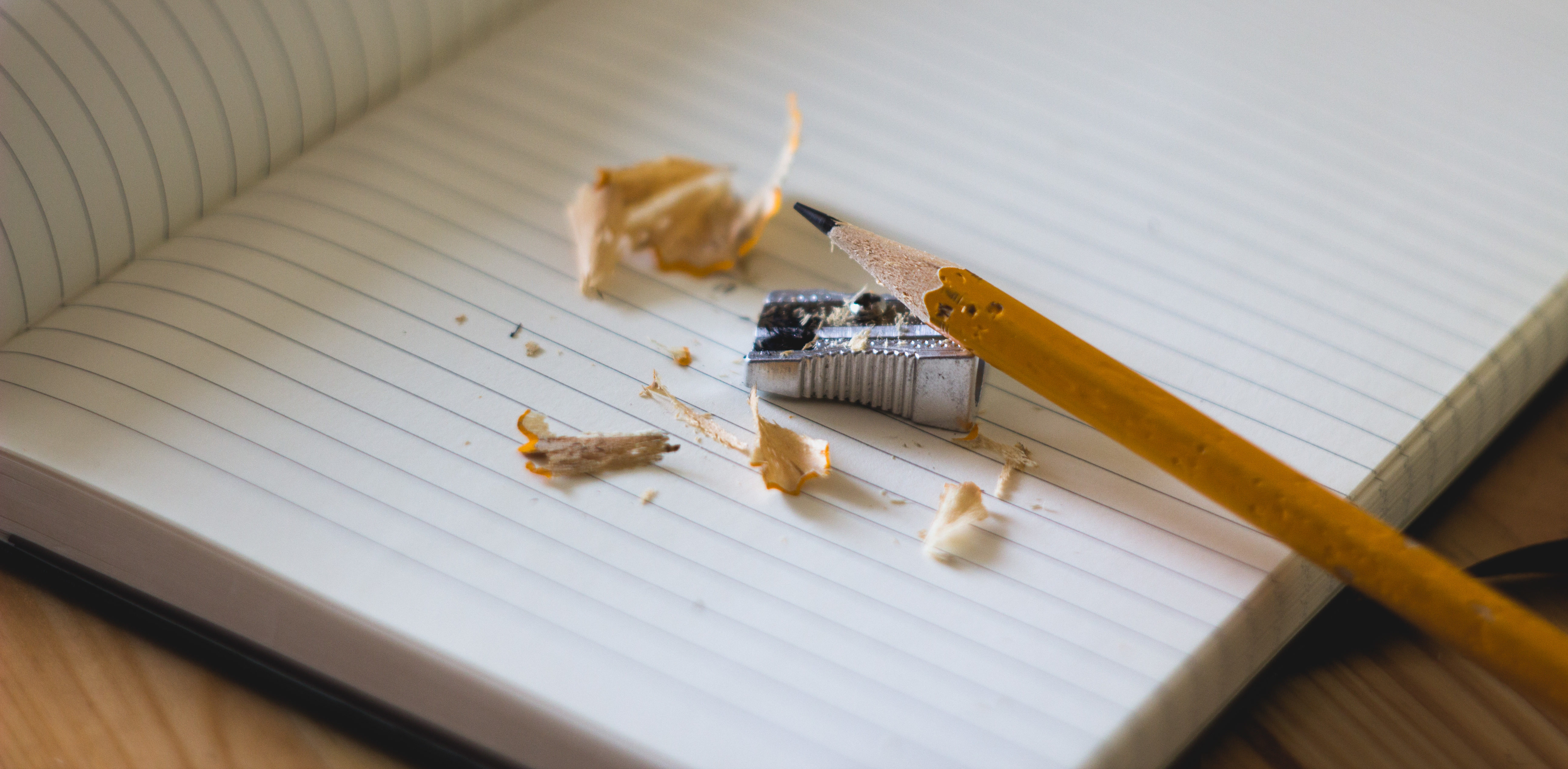 The image depicts a pencil and handheld sharpener on top of a blank, ruled notebook with pencil shavings around the sharpener.