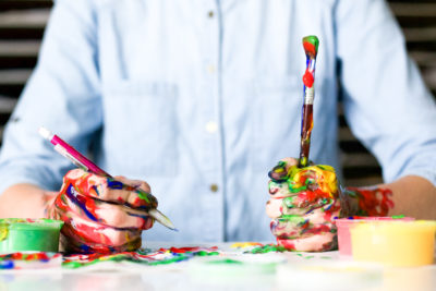 The image depicts a Caucasian person in a button-up shirt holding a paintbrush in one hand and a pencil in the other. Their hands and work surface are covered in many colors of paint.
