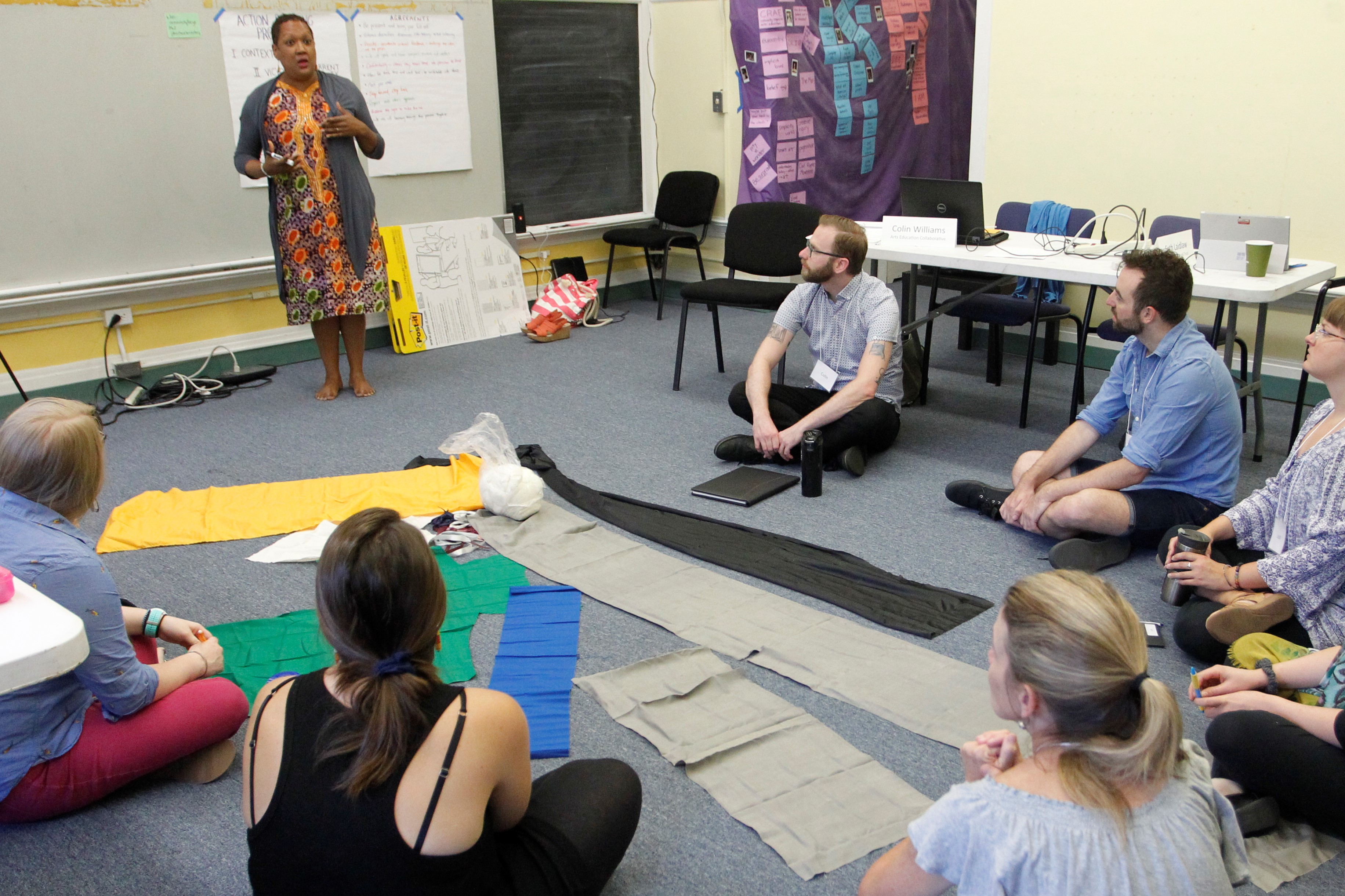 An educator facilitates an activity using colorful cloth with participants sitting on the floor in a circle.