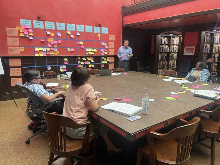 Three people listening to a evaluation services facilitator, several sticky notes and papers mapped out on the wall