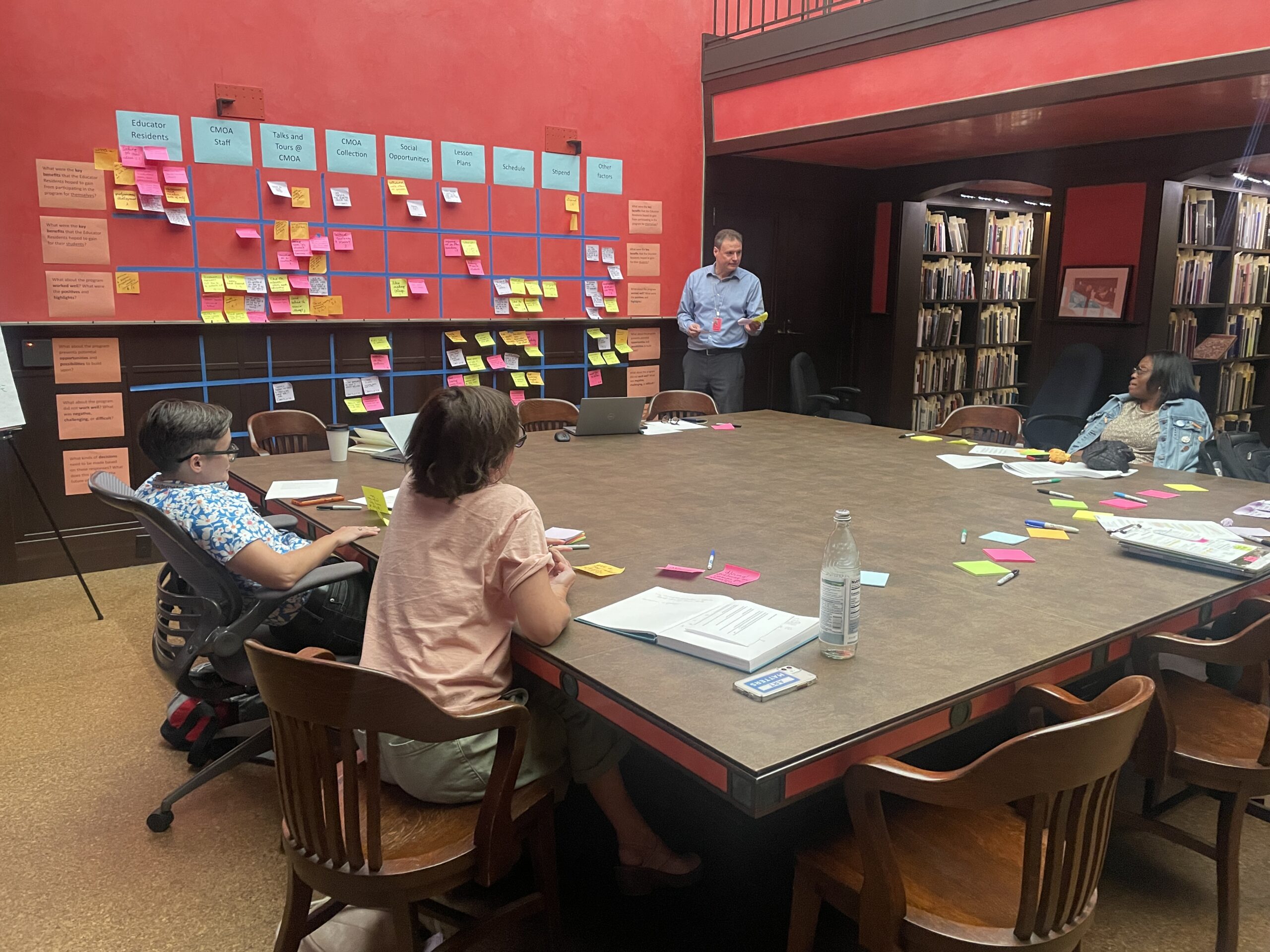 Three people listening to a evaluation services facilitator, several sticky notes and papers mapped out on the wall